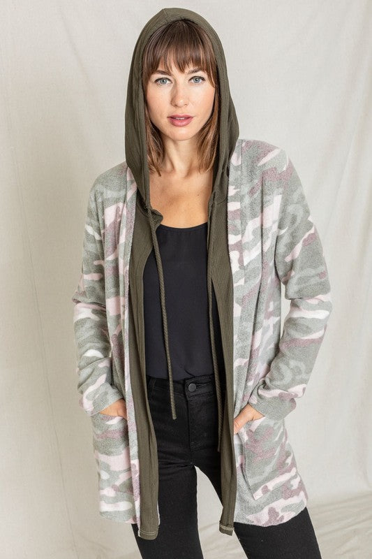 A woman wears a Camo Cardigan with a hoodie over a white top and jeans, standing gracefully against a pale backdrop, effortlessly blending casual style with modern flair.