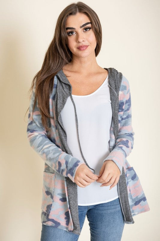 A woman wears a Camo Cardigan with a hoodie over a white top and jeans, standing gracefully against a pale backdrop, effortlessly blending casual style with modern flair.