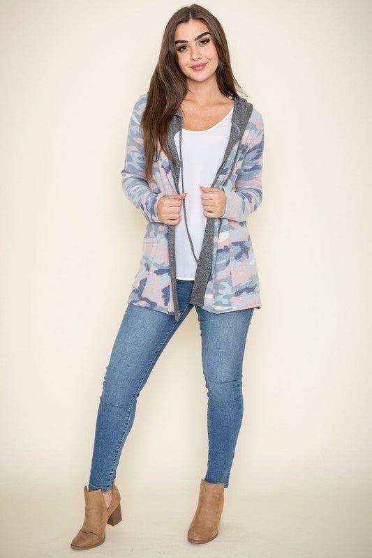 A woman wears a Camo Cardigan with a hoodie over a white top and jeans, standing gracefully against a pale backdrop, effortlessly blending casual style with modern flair.