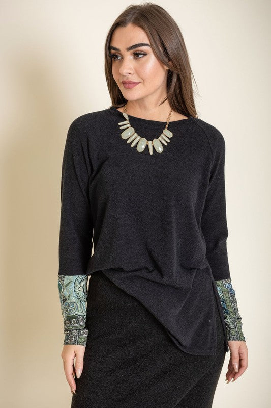 A person smiles with their eyes closed, wearing the Knit Side Slit Paisley Cuff Tunic in black, elegantly featuring paisley cuffs, with one hand touching their hair.