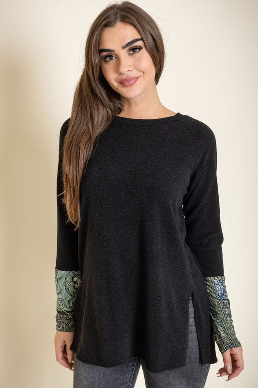 A person smiles with their eyes closed, wearing the Knit Side Slit Paisley Cuff Tunic in black, elegantly featuring paisley cuffs, with one hand touching their hair.