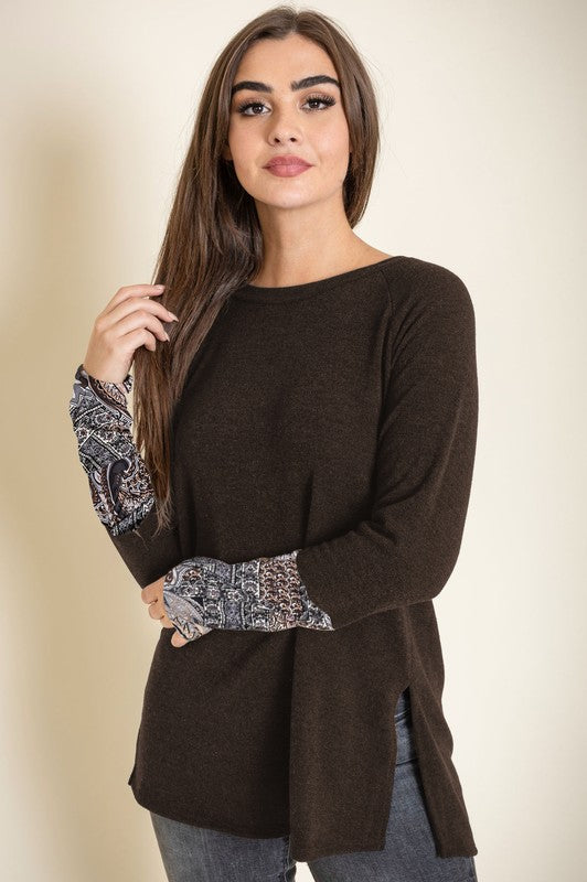 A person smiles with their eyes closed, wearing the Knit Side Slit Paisley Cuff Tunic in black, elegantly featuring paisley cuffs, with one hand touching their hair.