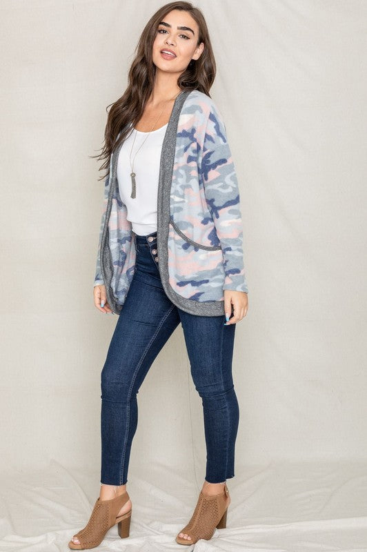 A woman is wearing the Camo Print Slouch Cardigan in a pink camo pattern with front pockets, styled with a white shirt and blue jeans.