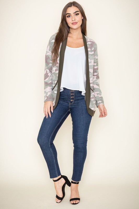 A woman is wearing the Camo Print Slouch Cardigan in a pink camo pattern with front pockets, styled with a white shirt and blue jeans.