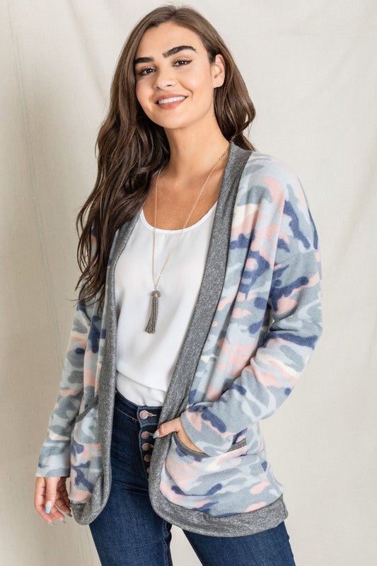 A woman is wearing the Camo Print Slouch Cardigan in a pink camo pattern with front pockets, styled with a white shirt and blue jeans.