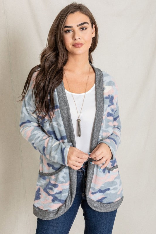 A woman is wearing the Camo Print Slouch Cardigan in a pink camo pattern with front pockets, styled with a white shirt and blue jeans.