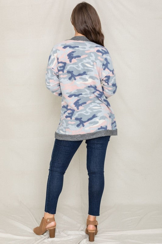 A woman is wearing the Camo Print Slouch Cardigan in a pink camo pattern with front pockets, styled with a white shirt and blue jeans.