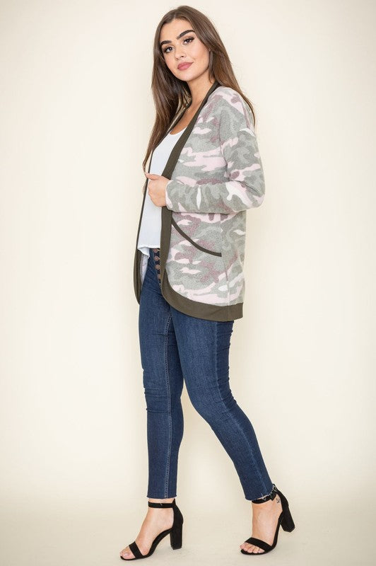 A woman is wearing the Camo Print Slouch Cardigan in a pink camo pattern with front pockets, styled with a white shirt and blue jeans.