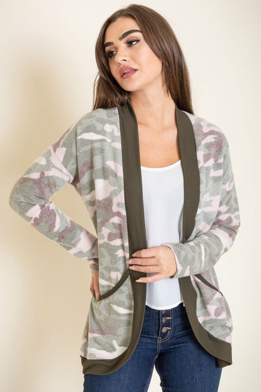 A woman is wearing the Camo Print Slouch Cardigan in a pink camo pattern with front pockets, styled with a white shirt and blue jeans.