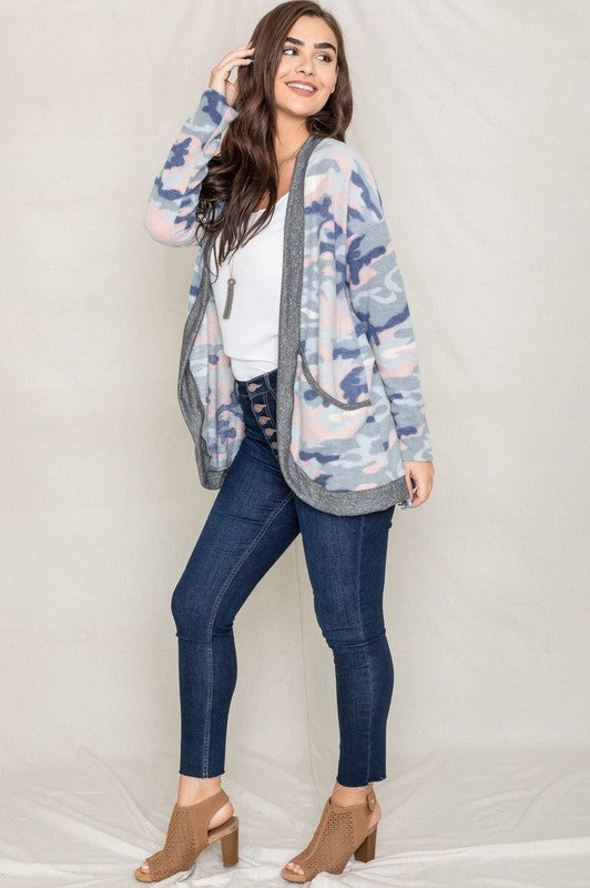 A woman is wearing the Camo Print Slouch Cardigan in a pink camo pattern with front pockets, styled with a white shirt and blue jeans.