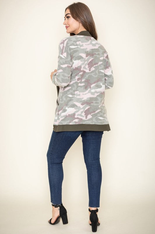 A woman is wearing the Camo Print Slouch Cardigan in a pink camo pattern with front pockets, styled with a white shirt and blue jeans.