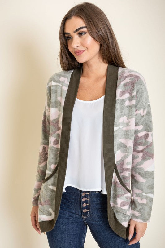 A woman is wearing the Camo Print Slouch Cardigan in a pink camo pattern with front pockets, styled with a white shirt and blue jeans.