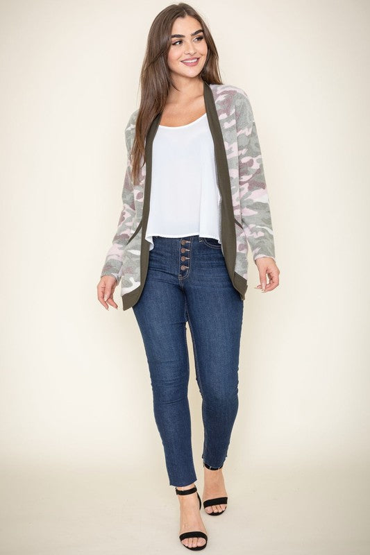 A woman is wearing the Camo Print Slouch Cardigan in a pink camo pattern with front pockets, styled with a white shirt and blue jeans.