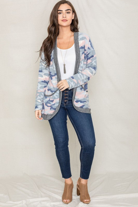 A woman is wearing the Camo Print Slouch Cardigan in a pink camo pattern with front pockets, styled with a white shirt and blue jeans.