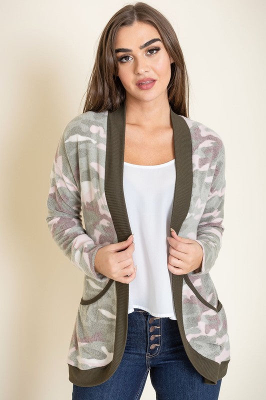 A woman is wearing the Camo Print Slouch Cardigan in a pink camo pattern with front pockets, styled with a white shirt and blue jeans.