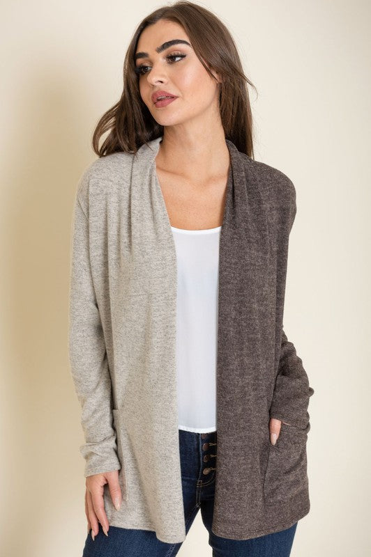 A woman proudly wearing the Two Tone Knit Cardigan, paired with a black top and jeans, smiles as she stands with her hand touching her hair. She accessorizes with a long necklace featuring a pendant, all crafted in the United States.