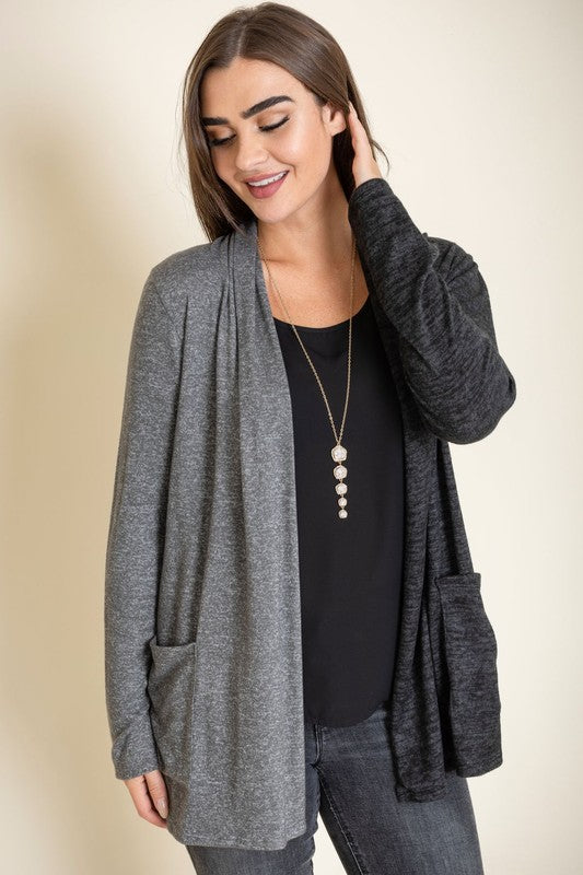 A woman proudly wearing the Two Tone Knit Cardigan, paired with a black top and jeans, smiles as she stands with her hand touching her hair. She accessorizes with a long necklace featuring a pendant, all crafted in the United States.