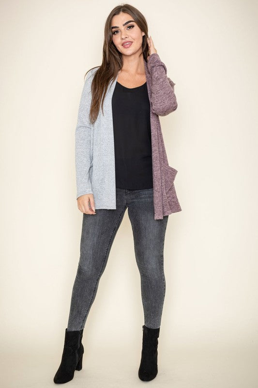 A woman proudly wearing the Two Tone Knit Cardigan, paired with a black top and jeans, smiles as she stands with her hand touching her hair. She accessorizes with a long necklace featuring a pendant, all crafted in the United States.