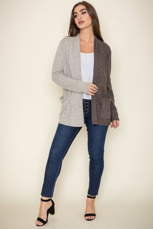 A woman proudly wearing the Two Tone Knit Cardigan, paired with a black top and jeans, smiles as she stands with her hand touching her hair. She accessorizes with a long necklace featuring a pendant, all crafted in the United States.