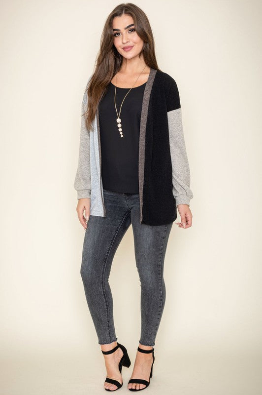 A woman is smiling and touching her hair while wearing the Color Block Knit Cardigan, featuring shades of black, grey, and beige. She has paired it with a black top and grey jeans. The cardigan is made from 95% rayon and is proudly manufactured in the United States.