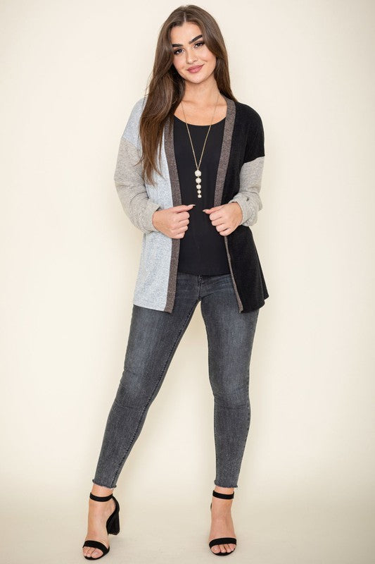 A woman is smiling and touching her hair while wearing the Color Block Knit Cardigan, featuring shades of black, grey, and beige. She has paired it with a black top and grey jeans. The cardigan is made from 95% rayon and is proudly manufactured in the United States.