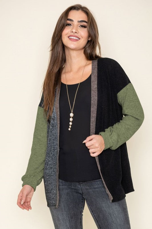 A woman with long brown hair wears a black top, the Color Block Knit Cardigan featuring green sleeves, and a necklace adorned with pearl-like beads while smiling.