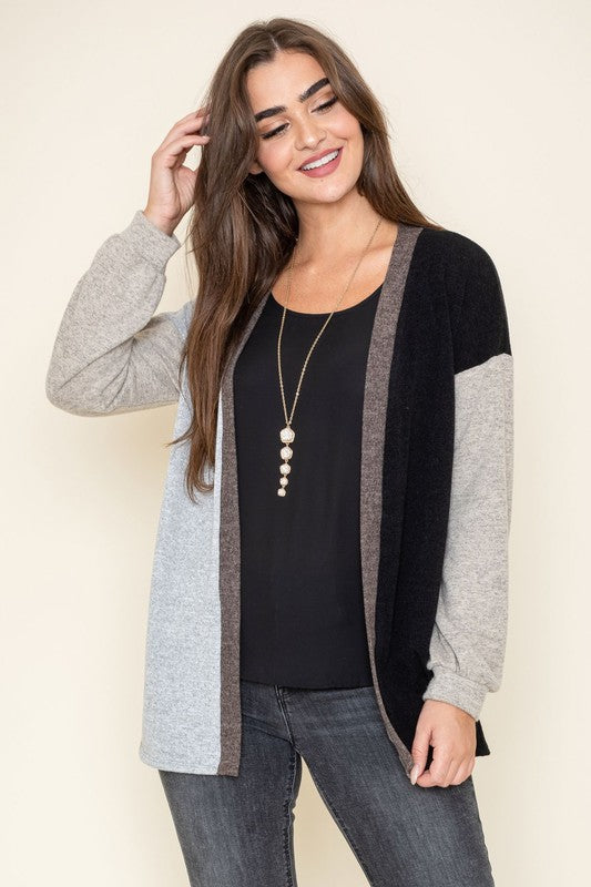 A woman is smiling and touching her hair while wearing the Color Block Knit Cardigan, featuring shades of black, grey, and beige. She has paired it with a black top and grey jeans. The cardigan is made from 95% rayon and is proudly manufactured in the United States.