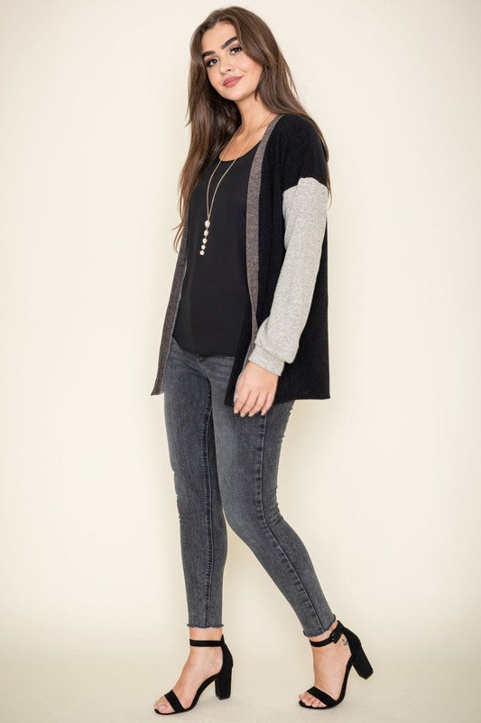 A woman is smiling and touching her hair while wearing the Color Block Knit Cardigan, featuring shades of black, grey, and beige. She has paired it with a black top and grey jeans. The cardigan is made from 95% rayon and is proudly manufactured in the United States.