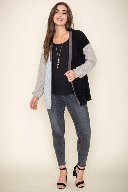 A woman is smiling and touching her hair while wearing the Color Block Knit Cardigan, featuring shades of black, grey, and beige. She has paired it with a black top and grey jeans. The cardigan is made from 95% rayon and is proudly manufactured in the United States.