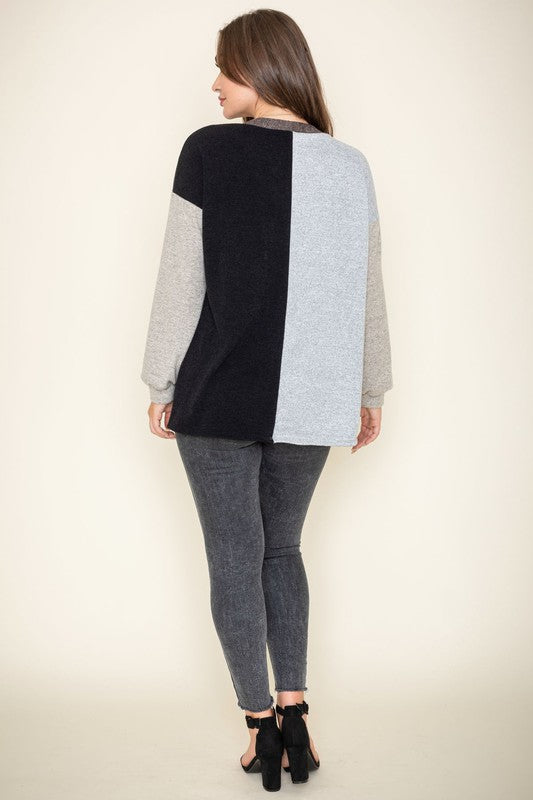 A woman is smiling and touching her hair while wearing the Color Block Knit Cardigan, featuring shades of black, grey, and beige. She has paired it with a black top and grey jeans. The cardigan is made from 95% rayon and is proudly manufactured in the United States.