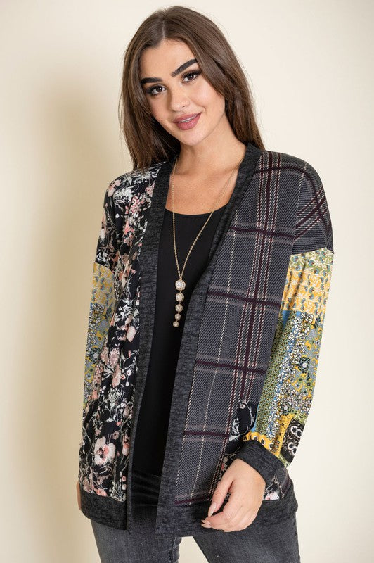 A woman wearing the Mix Fabric Open Cardigan, featuring floral and plaid designs, stands against a plain background, smiling. Crafted in the United States, this patchwork-patterned piece beautifully combines a variety of textures and prints.