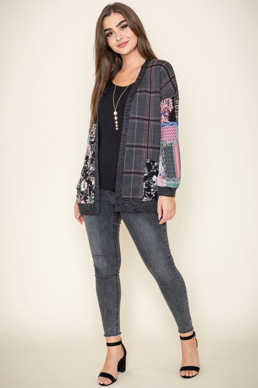 A woman wearing the Mix Fabric Open Cardigan, featuring floral and plaid designs, stands against a plain background, smiling. Crafted in the United States, this patchwork-patterned piece beautifully combines a variety of textures and prints.