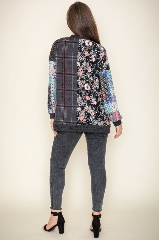 A woman wearing the Mix Fabric Open Cardigan, featuring floral and plaid designs, stands against a plain background, smiling. Crafted in the United States, this patchwork-patterned piece beautifully combines a variety of textures and prints.