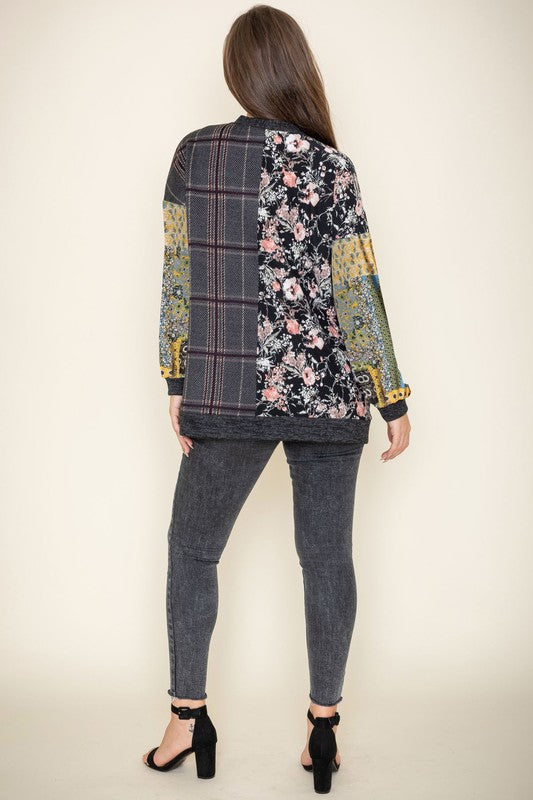 A woman wearing the Mix Fabric Open Cardigan, featuring floral and plaid designs, stands against a plain background, smiling. Crafted in the United States, this patchwork-patterned piece beautifully combines a variety of textures and prints.