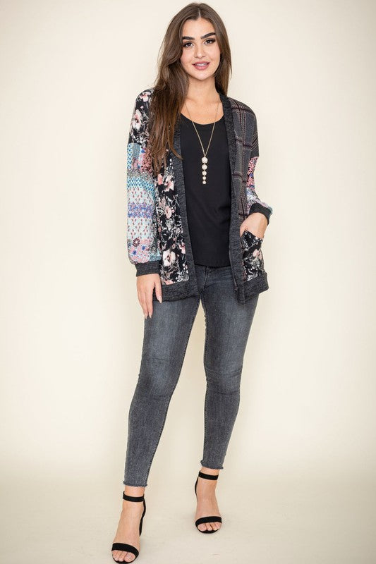 A woman wearing the Mix Fabric Open Cardigan, featuring floral and plaid designs, stands against a plain background, smiling. Crafted in the United States, this patchwork-patterned piece beautifully combines a variety of textures and prints.