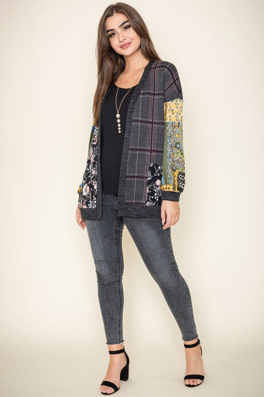 A woman wearing the Mix Fabric Open Cardigan, featuring floral and plaid designs, stands against a plain background, smiling. Crafted in the United States, this patchwork-patterned piece beautifully combines a variety of textures and prints.