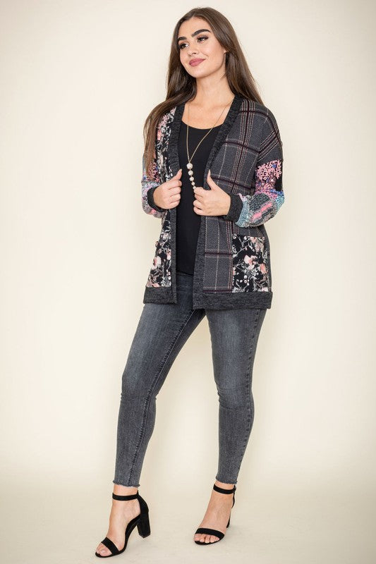 A woman wearing the Mix Fabric Open Cardigan, featuring floral and plaid designs, stands against a plain background, smiling. Crafted in the United States, this patchwork-patterned piece beautifully combines a variety of textures and prints.