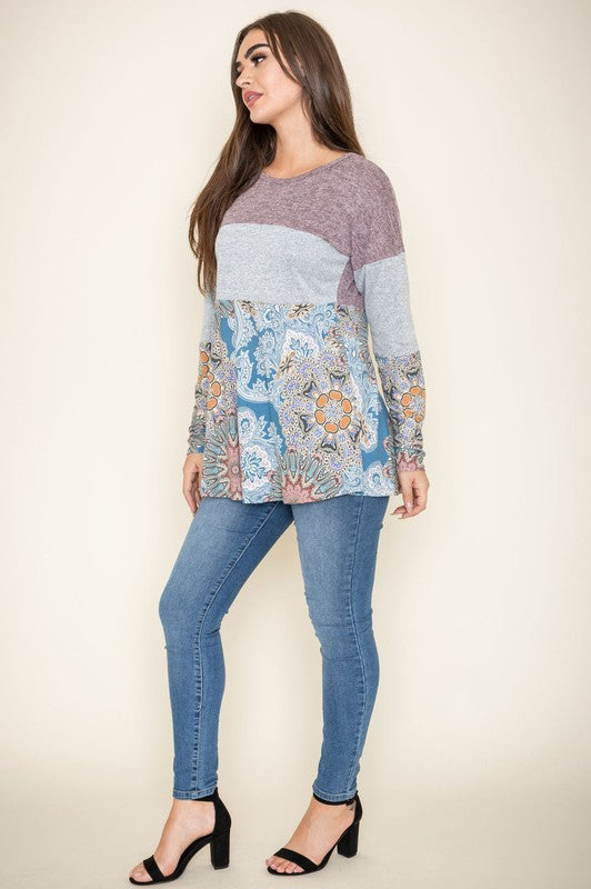 The woman stands gracefully in the Medallion Knit Color Block Tunic, paired with blue jeans and black heels. Her chic ensemble, crafted with a polyester spandex blend, highlights her style against the plain background.