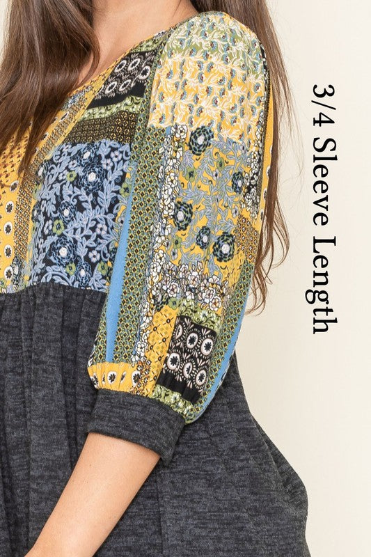 A woman with long brown hair wears the 3/4 Sleeve Quilted Pattern Tunic, featuring a colorful quilted pattern and a dark gray lower half. Made in the United States, she stands against a plain, light-colored background, smiling gently with her hands clasped.