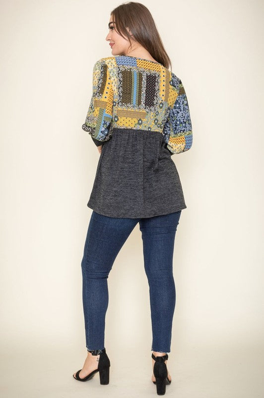 A woman with long brown hair wears the 3/4 Sleeve Quilted Pattern Tunic, featuring a colorful quilted pattern and a dark gray lower half. Made in the United States, she stands against a plain, light-colored background, smiling gently with her hands clasped.