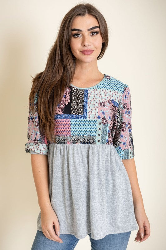 A woman with long brown hair wears the 3/4 Sleeve Quilted Pattern Tunic, featuring a colorful quilted pattern and a dark gray lower half. Made in the United States, she stands against a plain, light-colored background, smiling gently with her hands clasped.