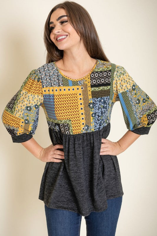 A woman with long brown hair wears the 3/4 Sleeve Quilted Pattern Tunic, featuring a colorful quilted pattern and a dark gray lower half. Made in the United States, she stands against a plain, light-colored background, smiling gently with her hands clasped.