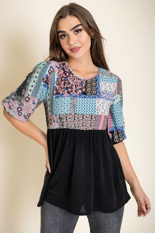 A woman with long brown hair wears the 3/4 Sleeve Quilted Pattern Tunic, featuring a colorful quilted pattern and a dark gray lower half. Made in the United States, she stands against a plain, light-colored background, smiling gently with her hands clasped.