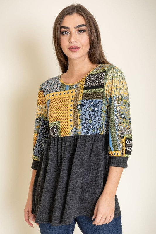 A woman with long brown hair wears the 3/4 Sleeve Quilted Pattern Tunic, featuring a colorful quilted pattern and a dark gray lower half. Made in the United States, she stands against a plain, light-colored background, smiling gently with her hands clasped.