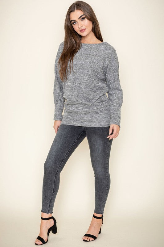 A woman with long brown hair wearing a gray knit sweater and dark jeans poses with one hand on her collarbone. This elegant Plus Tweed Blouson Tunic is of limited quantity, showcasing timeless fashion.