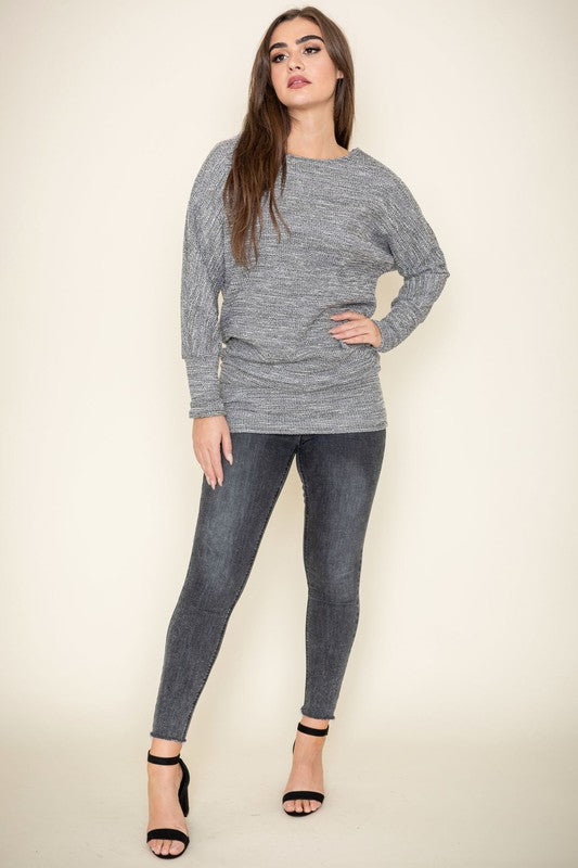 A woman with long brown hair wearing a gray knit sweater and dark jeans poses with one hand on her collarbone. This elegant Plus Tweed Blouson Tunic is of limited quantity, showcasing timeless fashion.