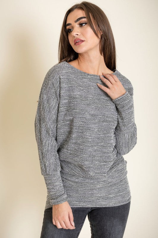 A woman with long brown hair wearing a grey Tweed Blouson Tunic and dark jeans stands against a plain background, touching her collarbone with her right hand. Limited quantity available; proudly made in the United States.