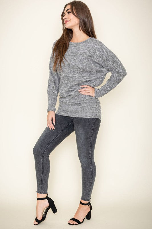 A woman with long brown hair wearing a gray knit sweater and dark jeans poses with one hand on her collarbone. This elegant Plus Tweed Blouson Tunic is of limited quantity, showcasing timeless fashion.