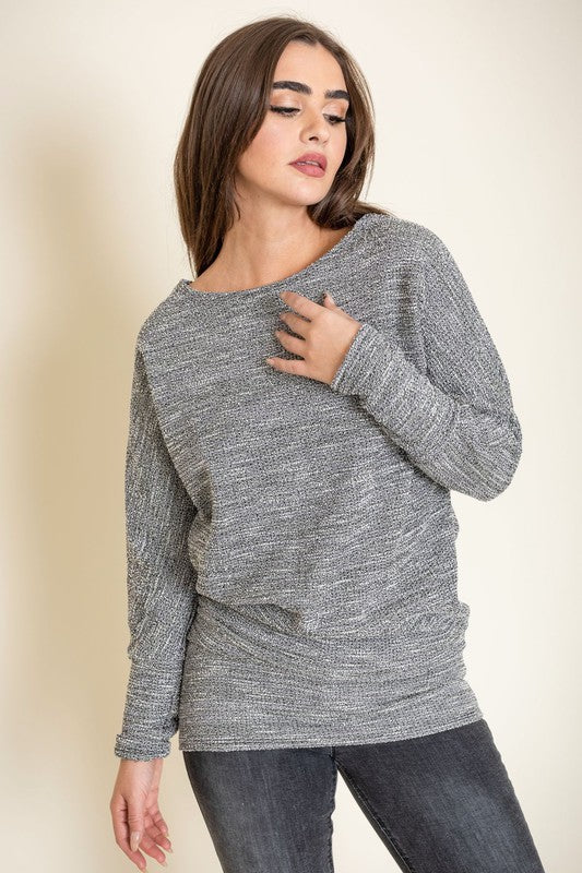 A woman with long brown hair wearing a gray knit sweater and dark jeans poses with one hand on her collarbone. This elegant Plus Tweed Blouson Tunic is of limited quantity, showcasing timeless fashion.