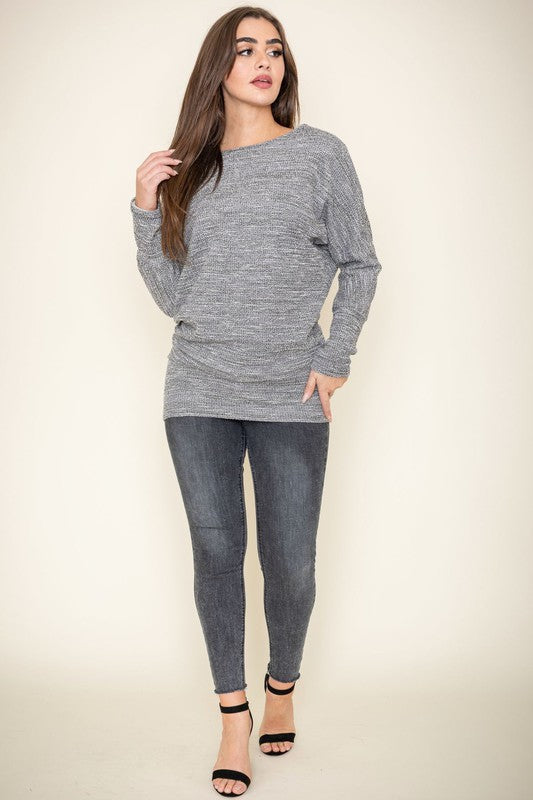 A woman with long brown hair wearing a gray knit sweater and dark jeans poses with one hand on her collarbone. This elegant Plus Tweed Blouson Tunic is of limited quantity, showcasing timeless fashion.